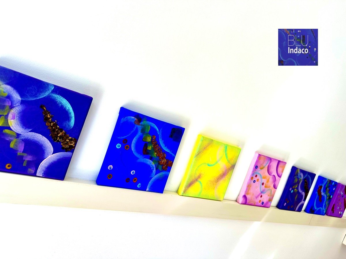 Sette tele di formati diversi appoggiate su una mensola bianca: blu indaco, giallo, rosa e viola, con cerchi chiari, punti colorati e inserti dorati.